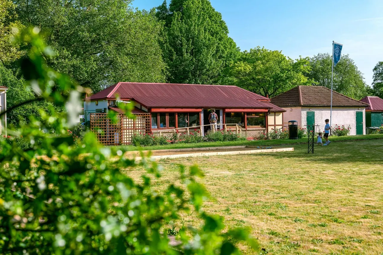 Low-angle view of a leisure venue building with red roof and terrace, surrounded by trees and green spaces, with visitors on the lawn.