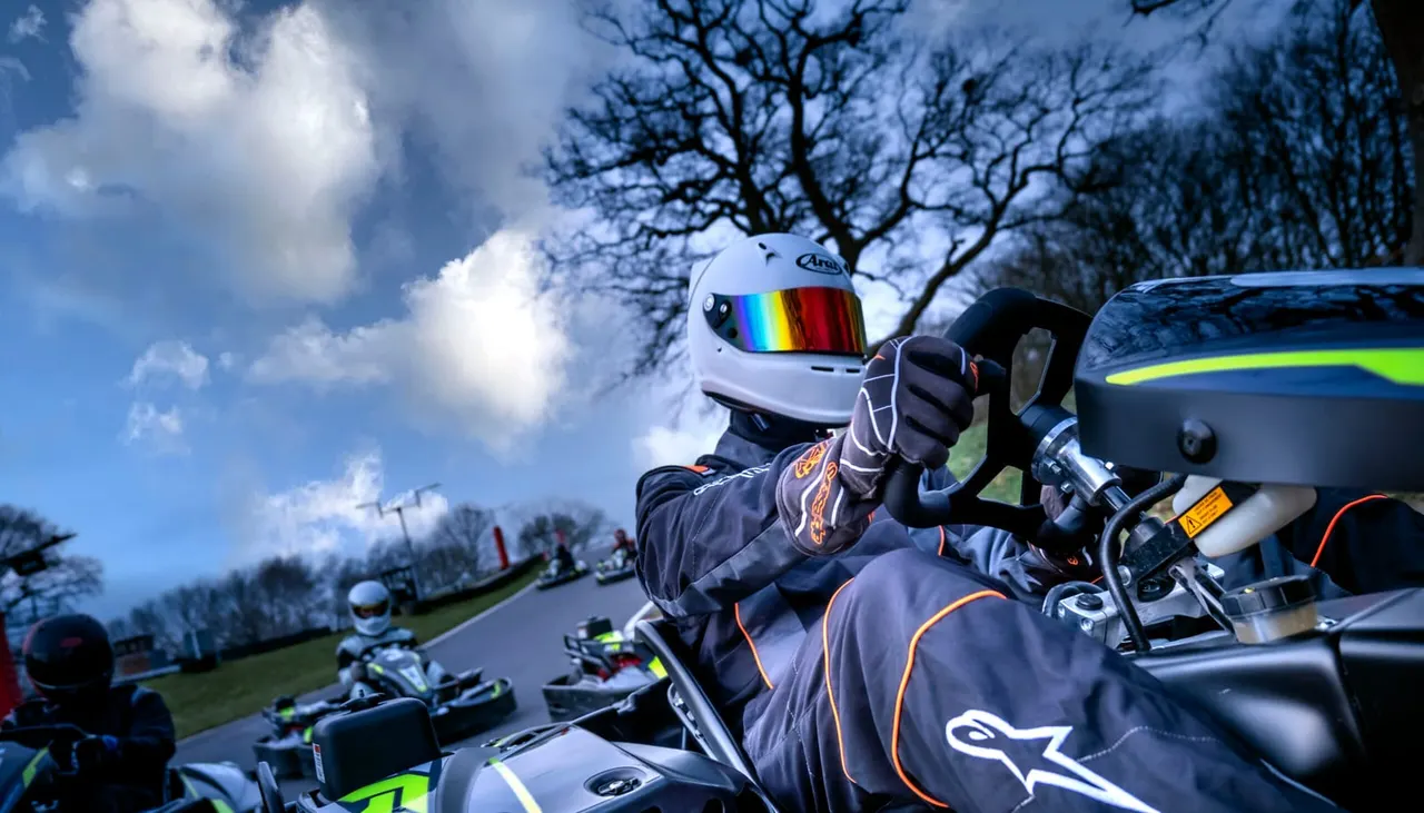 Go-kart racer in white helmet and dark racing suit gives thumbs up whilst seated in kart on outdoor track with other karts and spectators visible.