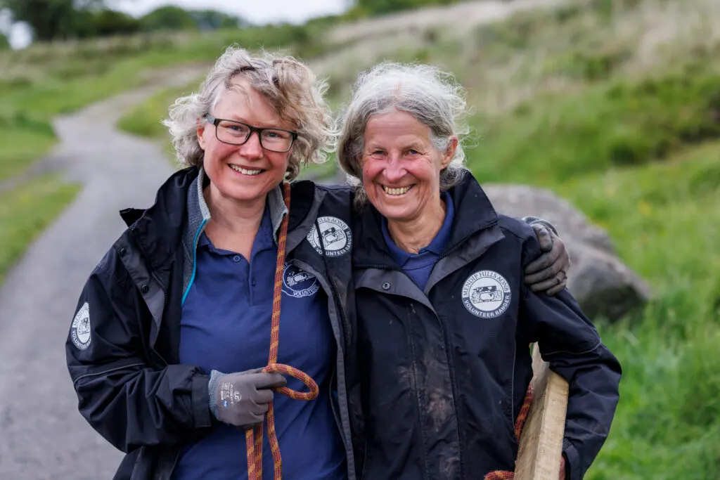 Two uniformed staff members smile whilst standing on a rural path with green hillsides behind them.