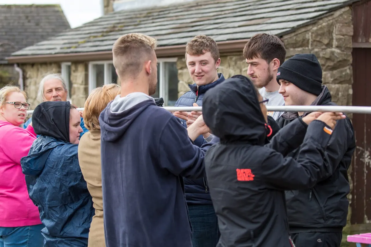 Group of visitors and staff gathered outdoors at a stone building, holding a metal pole together during a team activity.