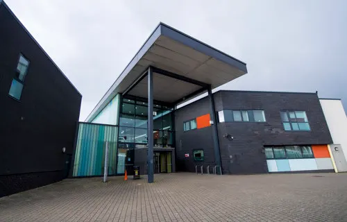 Modern single-storey building with dark brick exterior, glass entrance canopy and paved forecourt under cloudy sky.