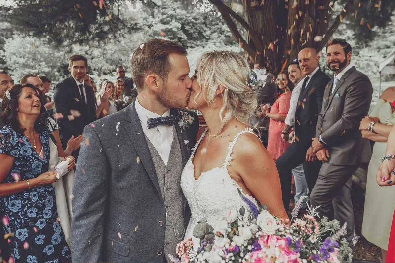 Bride and groom kiss during outdoor wedding ceremony under tree with guests gathered around.