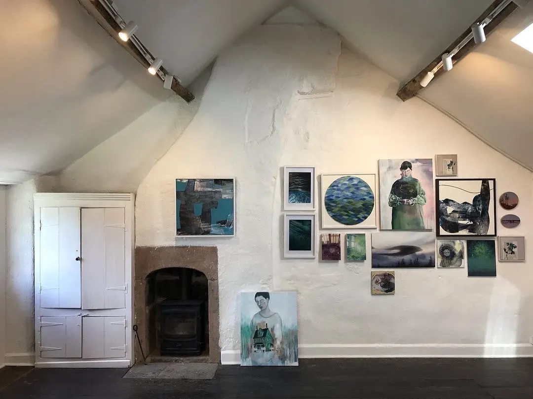 Interior of a gallery space with whitewashed walls, vaulted ceiling, fireplace, and various framed artworks and paintings displayed.