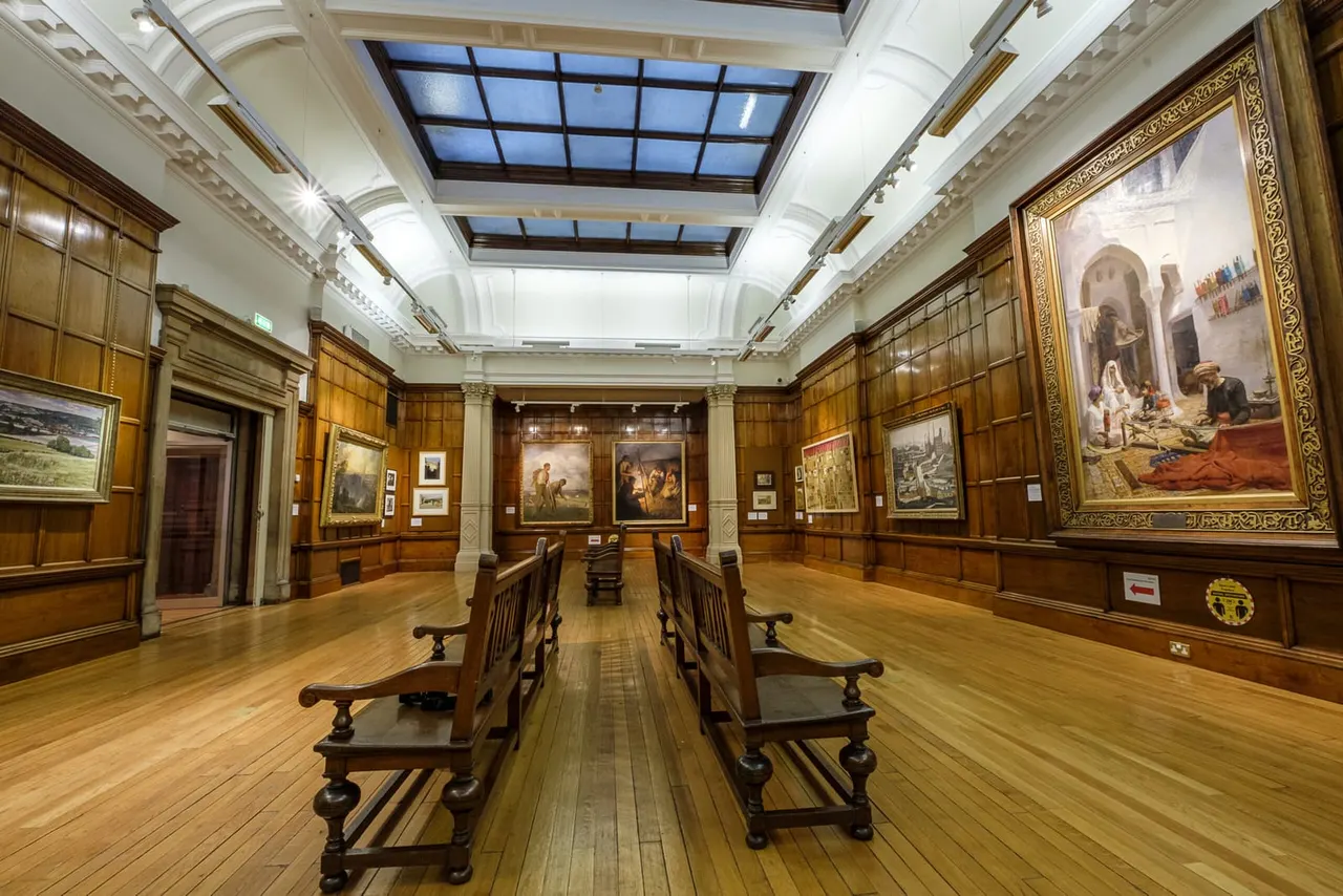 Empty gallery hall with wooden floors, panelled walls, skylights and framed paintings displayed on walls.