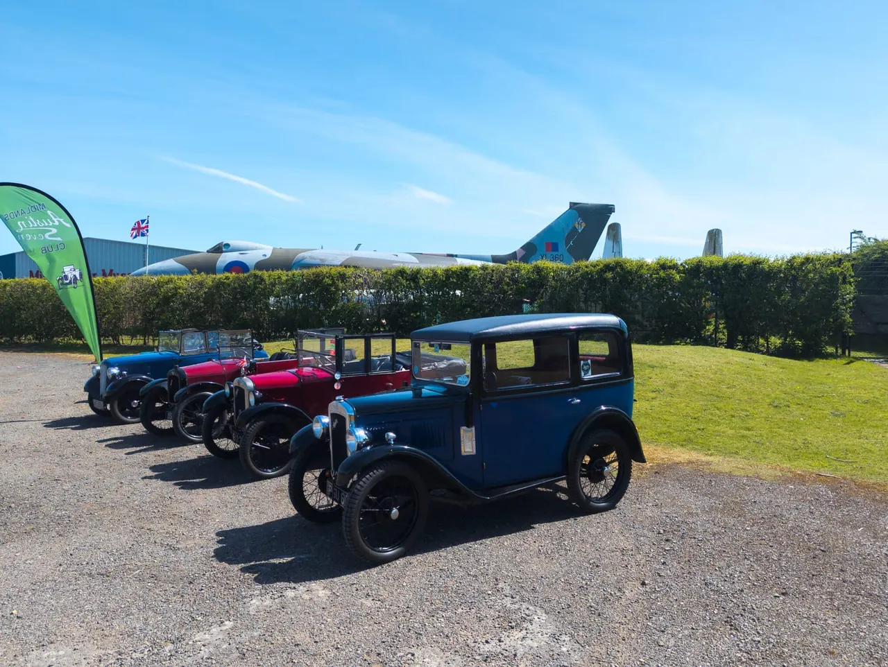 Vintage cars parked on a gravel area beside a hedge, with a decommissioned aircraft visible behind and clear blue sky above.