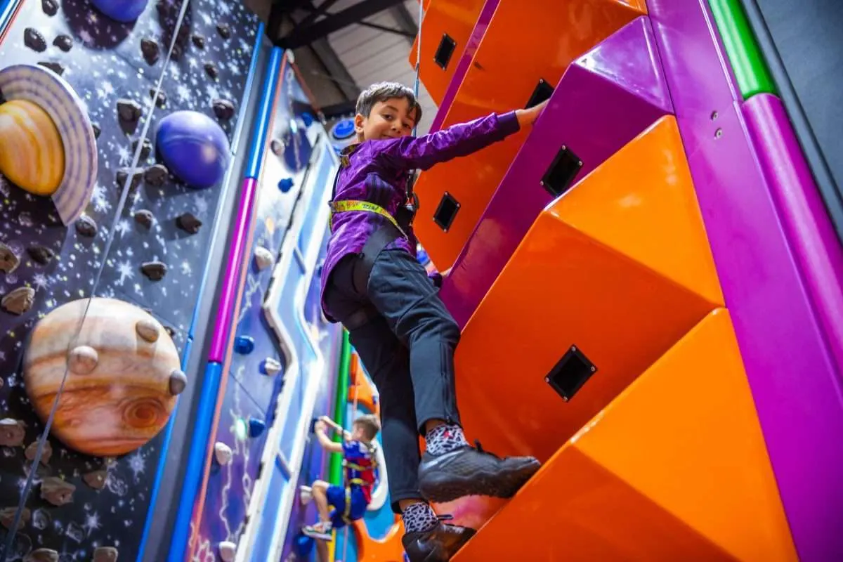 Child in purple jacket climbing indoor climbing wall with colourful holds and space-themed panels.