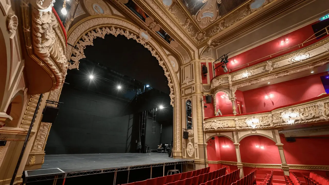 Historic theatre interior with ornate gilt ceiling, red velvet seats, and an empty stage framed by an elaborate proscenium arch.