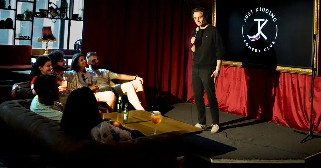 A comedian performs on stage at a comedy club whilst an audience of people sit on a sofa watching and holding drinks.