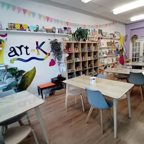 A bright art studio with work tables, shelving displaying artwork and supplies, colourful bunting, and an "art-K" mural on the wall.