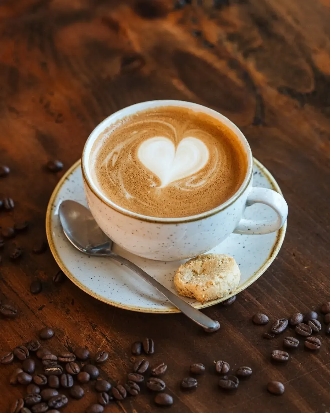 Cappuccino with latte art heart design in speckled white cup on saucer with biscuit and coffee beans on wooden table.