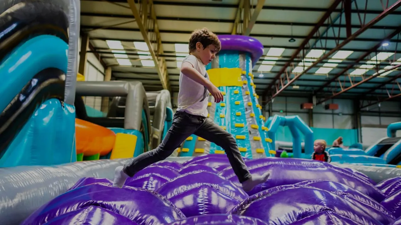Child jumping on purple inflatable obstacle course inside an indoor play facility with colourful inflatables and slides.