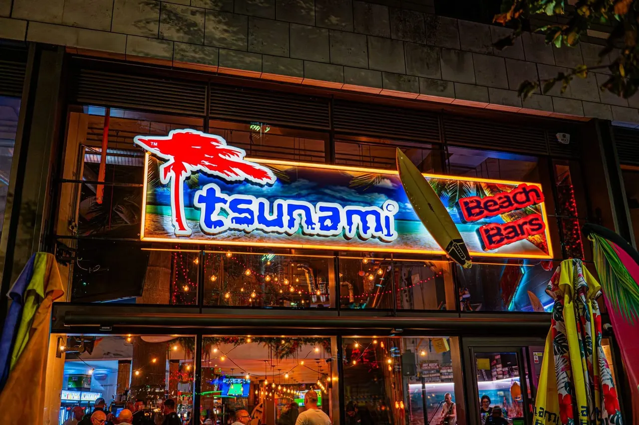 Neon-lit storefront of a beach bar at night, with illuminated red and blue signage, surfboards displayed outside, and people visible inside the lit venue.