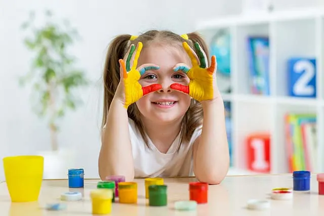 Child with paint-covered hands held up to face, smiling at table with paint pots and art supplies.