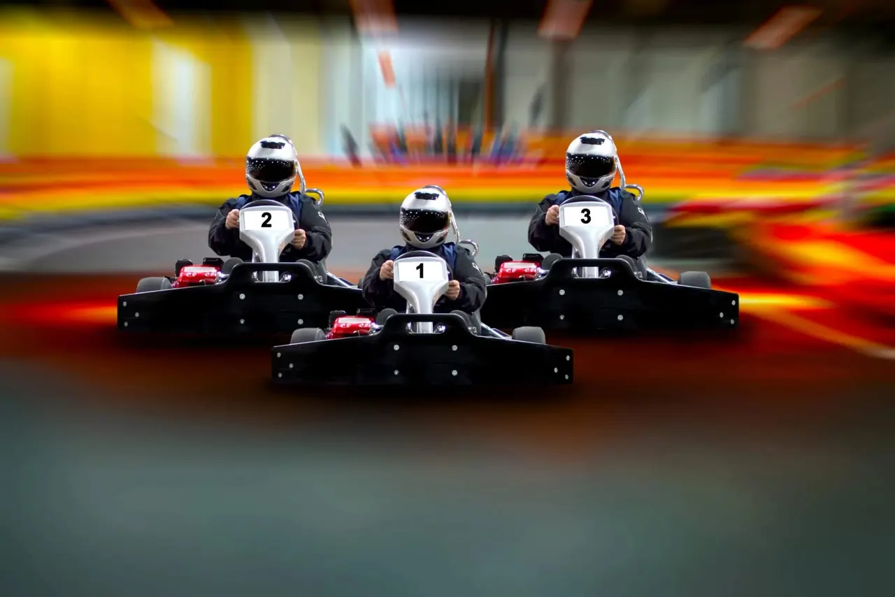 Three go-kart racers in helmets and numbered bibs driving on an indoor track with colourful barriers.