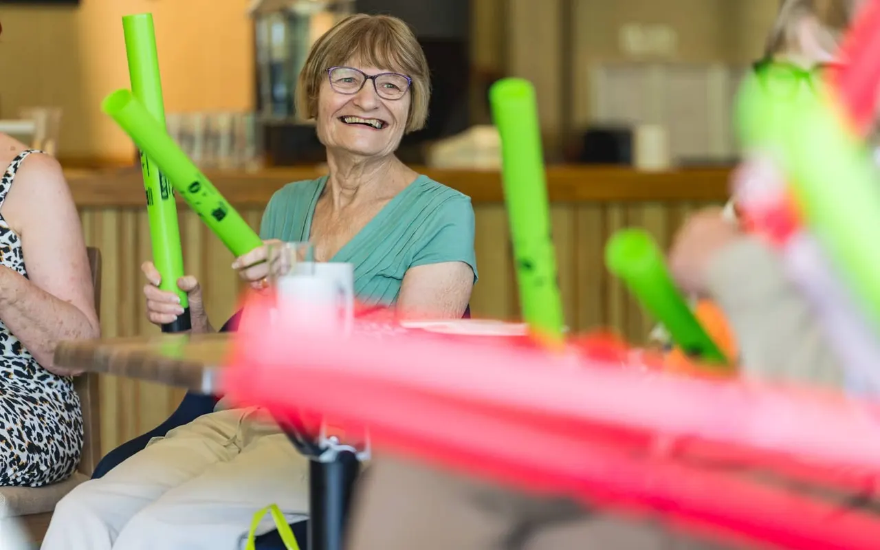 Older woman wearing glasses smiling while holding bright green foam nunchucks during an indoor activity session.