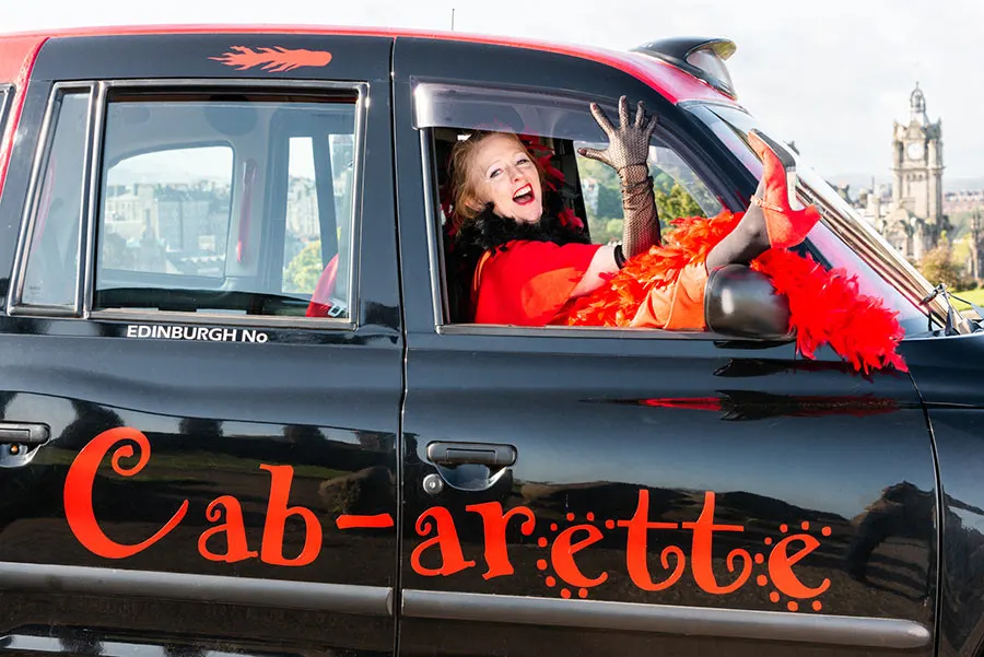 Woman in red outfit waving from window of black taxi cab decorated with red feathers, Edinburgh cityscape behind.