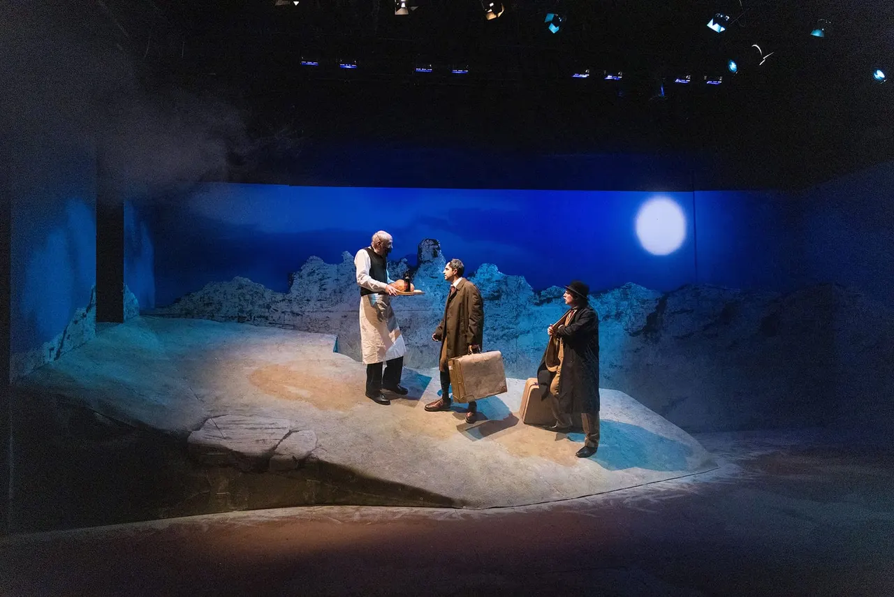 Actors perform a scene on an illuminated stage set with rocky terrain and blue lighting, featuring a full moon backdrop.