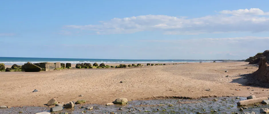 Sandy beach with shallow water in foreground, rock formations and groynes visible along shoreline stretching toward distant horizon under blue sky.