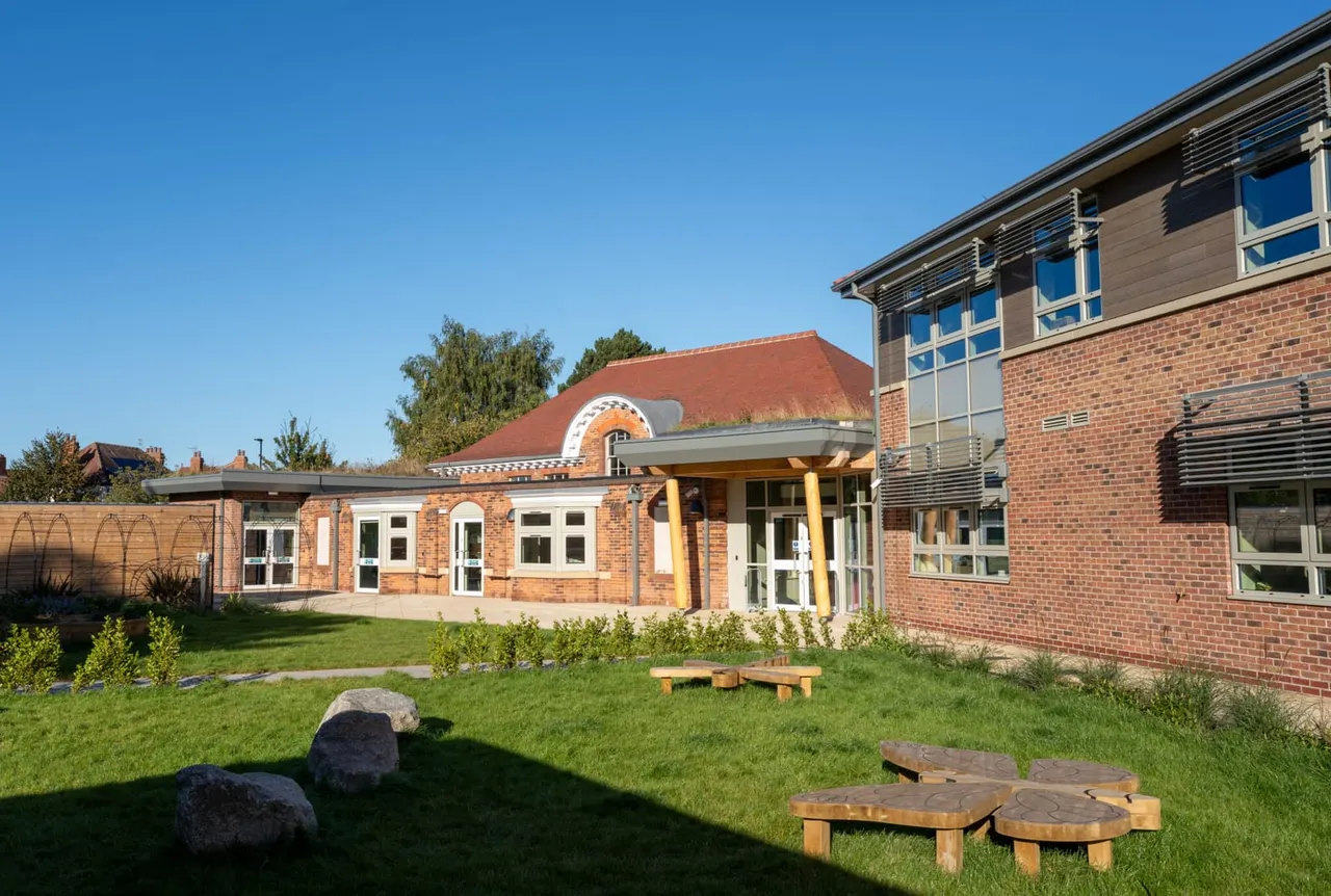 Modern brick leisure building with outdoor seating area featuring wooden benches and landscaped gardens under clear blue sky.