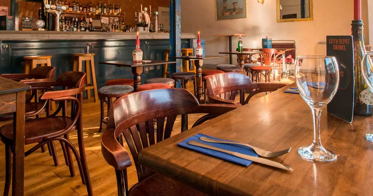 Warm-toned bar and dining room with wooden furniture, set tables, and a fully stocked bar counter in the background.