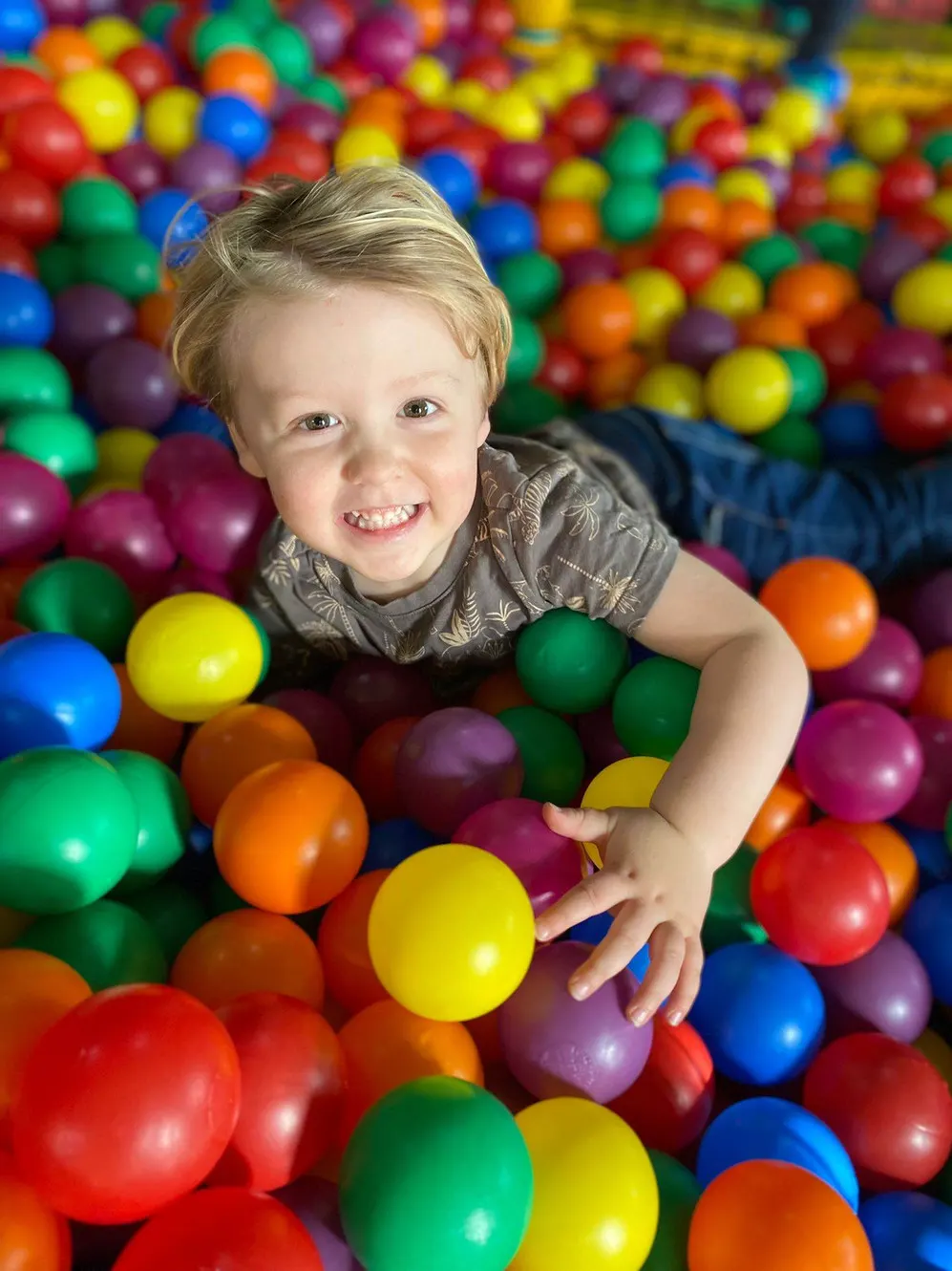 Young child smiling in a colourful ball pit filled with red, yellow, green, blue and orange balls.