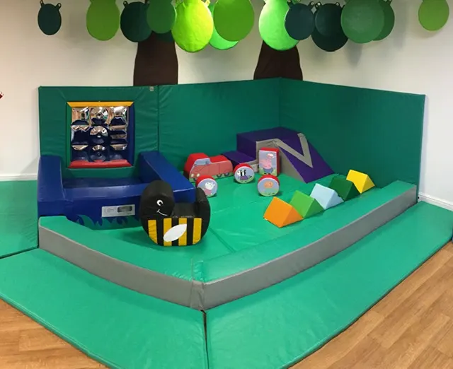 Soft play area with green padded surfaces, foam shapes, toy vehicles, and suspended green and black balloons for children's indoor play.