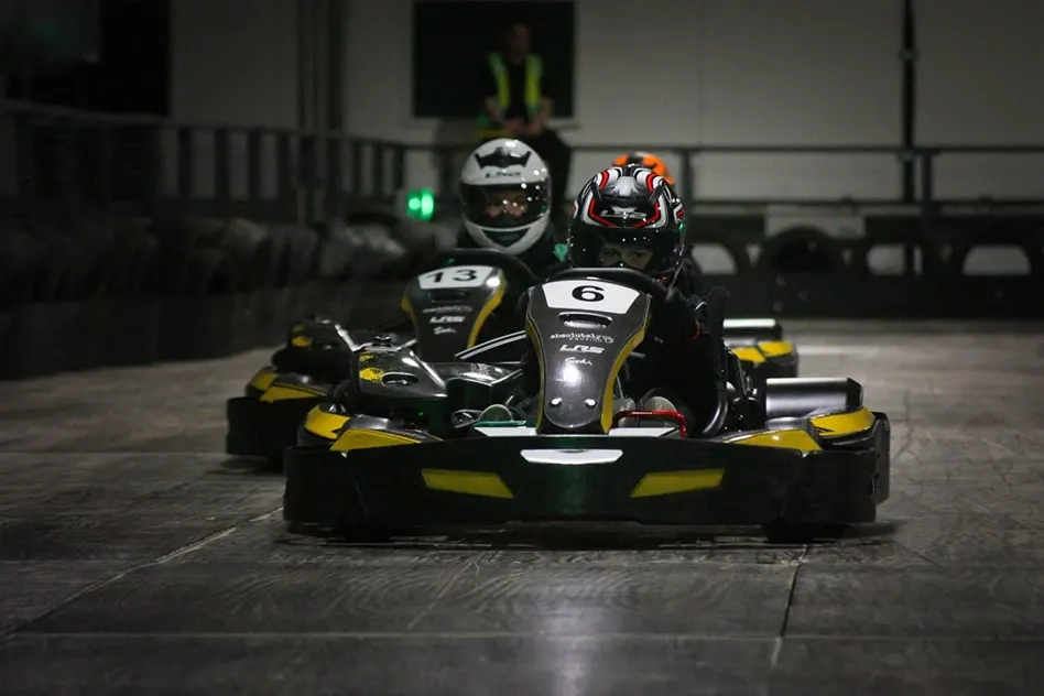 Two drivers in helmets racing go-karts indoors at night on a concrete track.
