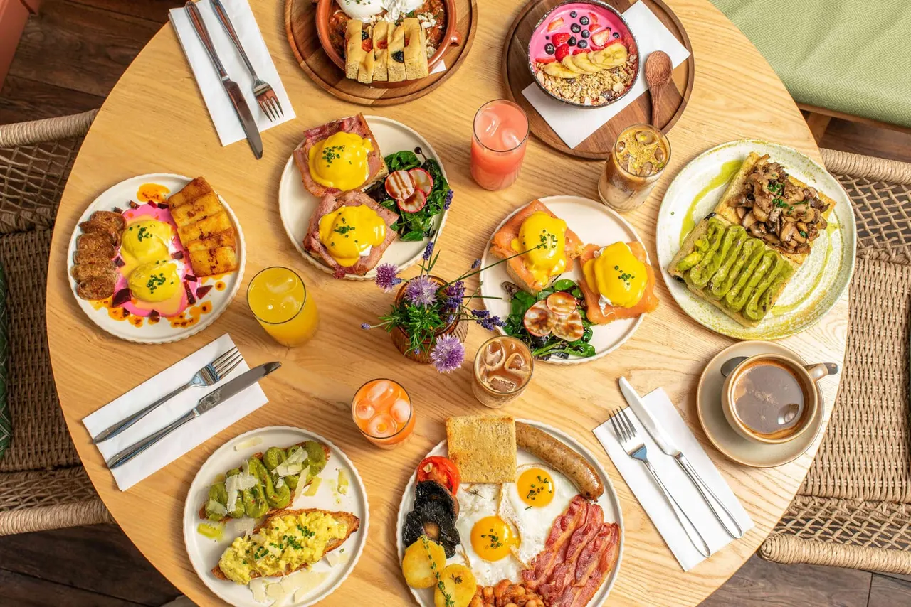 Overhead view of a round wooden table set for brunch with multiple plates of eggs, avocado toast, pastries, fresh juices and coffee.