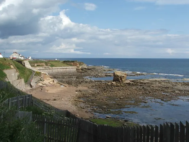 Coastal beach scene with rocky shoreline, sandy beach, and clifftop buildings under blue sky with white clouds.
