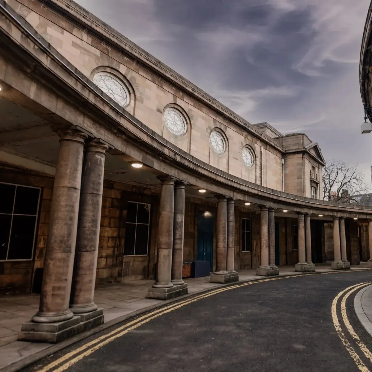 Historic stone building with classical columns and four circular clock faces on the façade, under a cloudy sky.