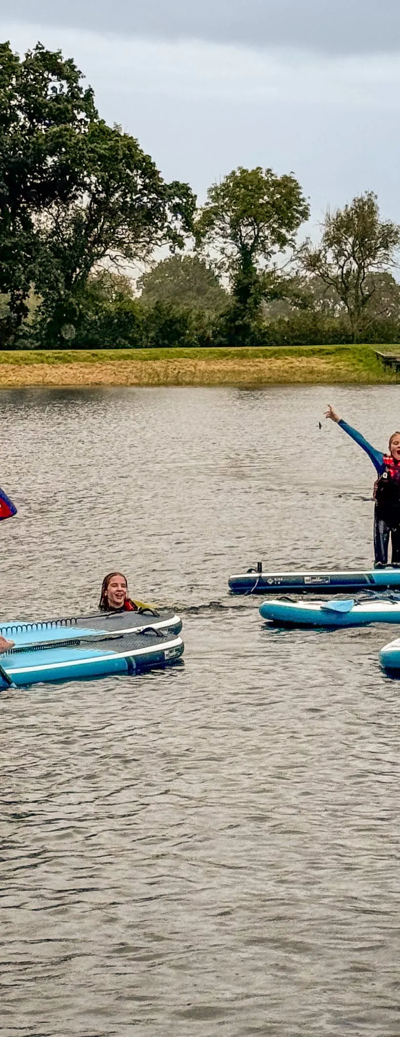 Children paddleboarding on a calm lake with trees lining the far bank under cloudy sky.
