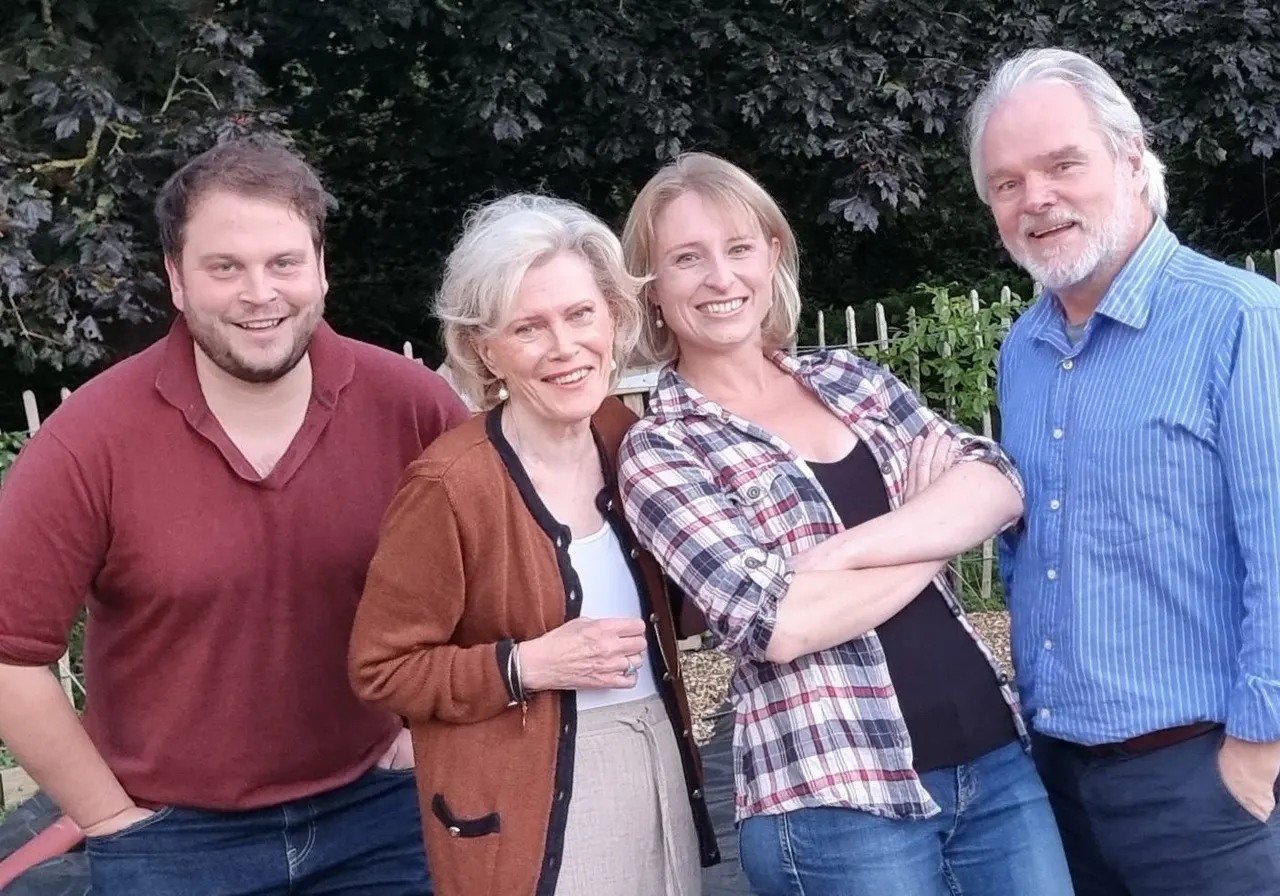 Four smiling adults posing together outdoors in front of dark foliage and a garden fence.