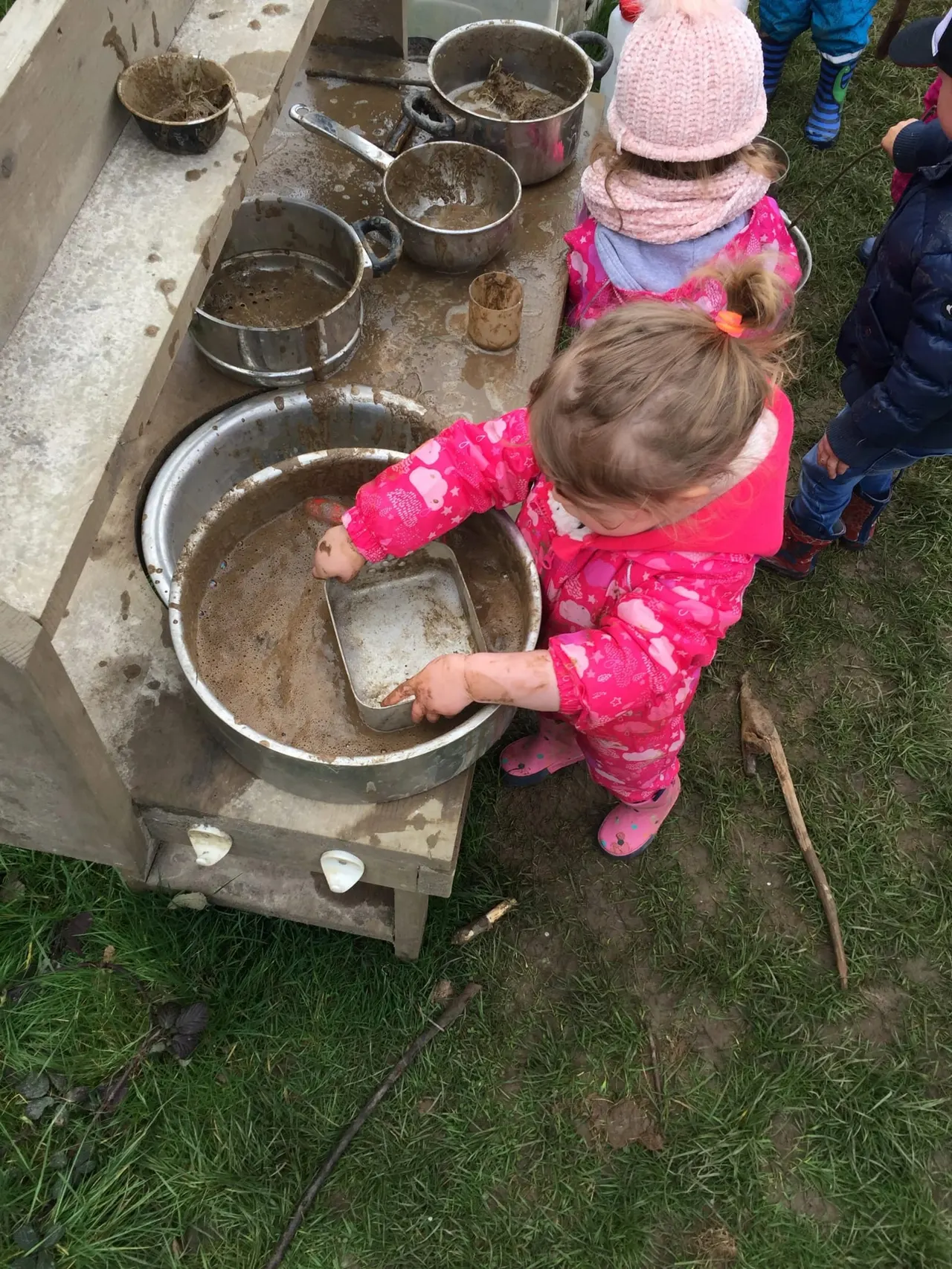 Young child in pink jacket and hat scooping mud from a large basin during an outdoor play activity with other children nearby.