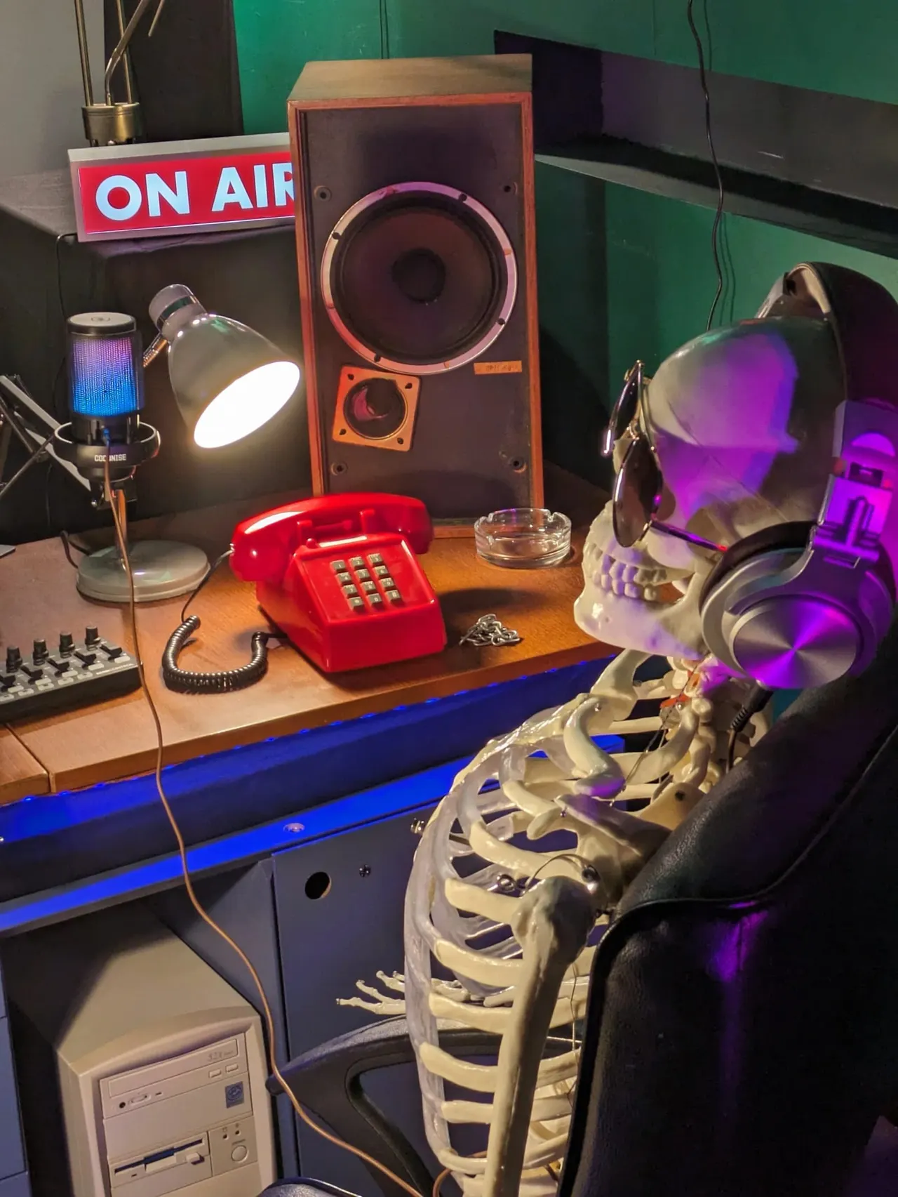 Radio studio set-up with skeleton mannequin wearing headphones at desk with vintage red telephone, microphone and speaker.