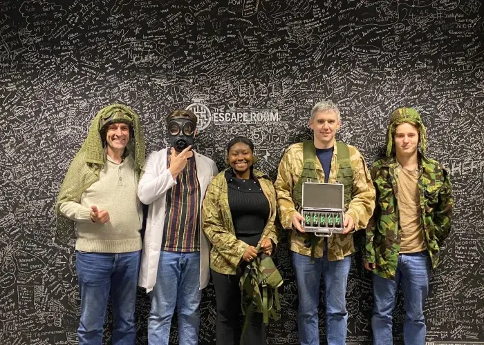 Five people in costume pose together against a wall covered in handwritten text and puzzle drawings.