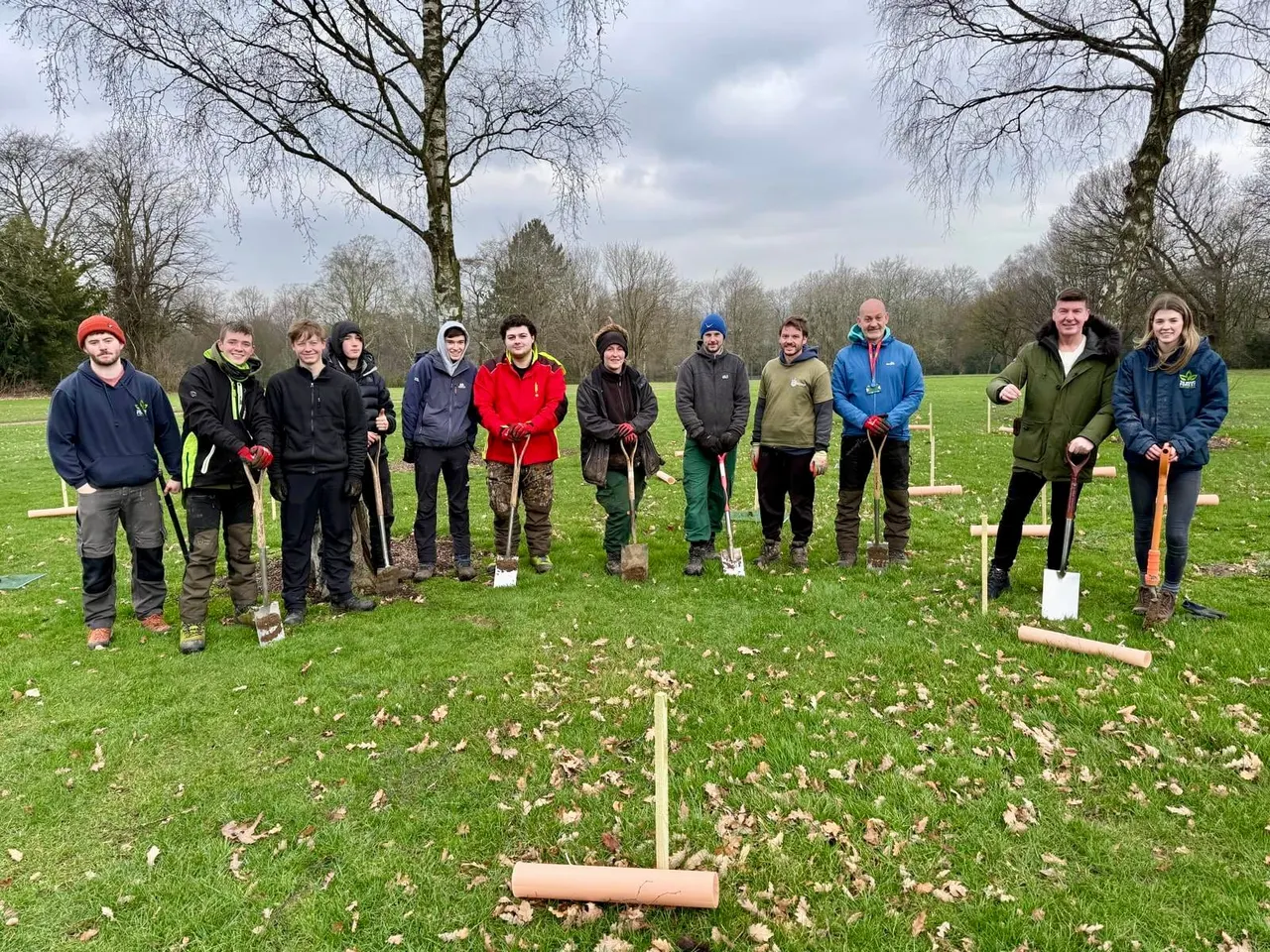 A group of twelve people holding spades stand in a line across a grassy field with bare trees, participating in a tree-planting activity.