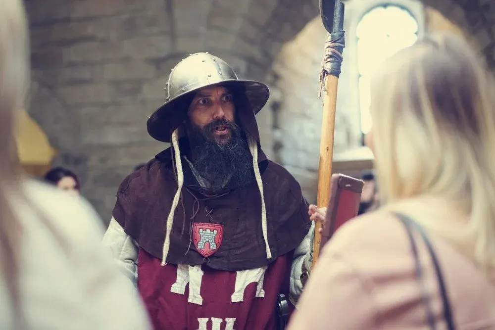 A bearded man dressed as a medieval warrior with helmet and axe speaks with a visitor inside a stone castle.