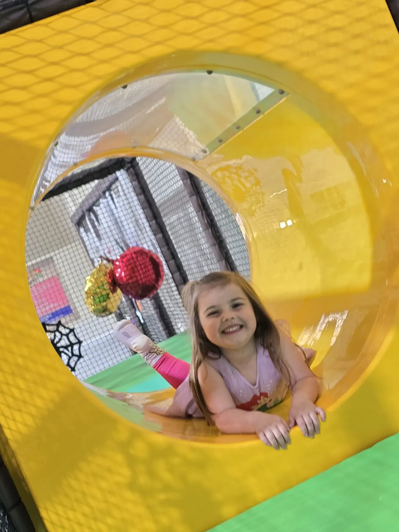 Young child smiling inside a yellow soft play tunnel with game targets visible through the opening.