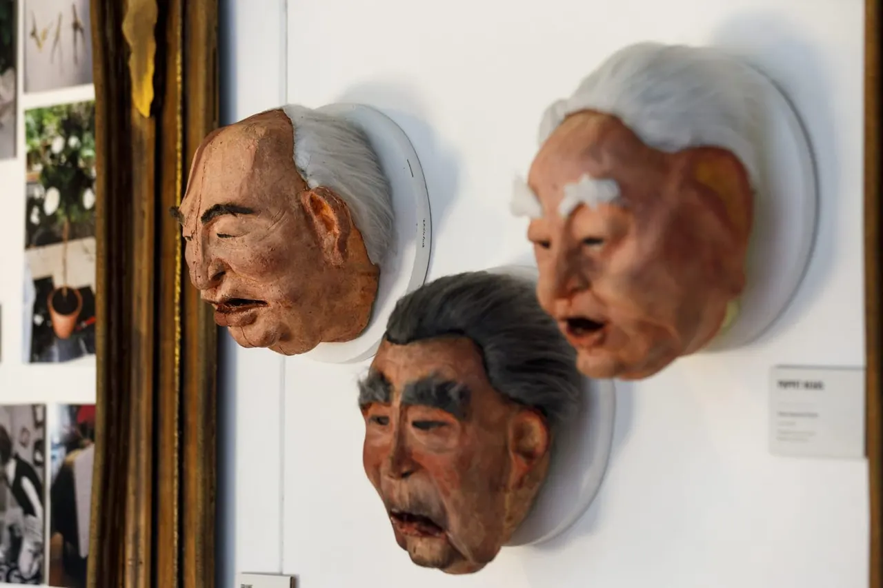 Three sculptured head masks mounted on a white gallery wall, with photographs visible to the left.