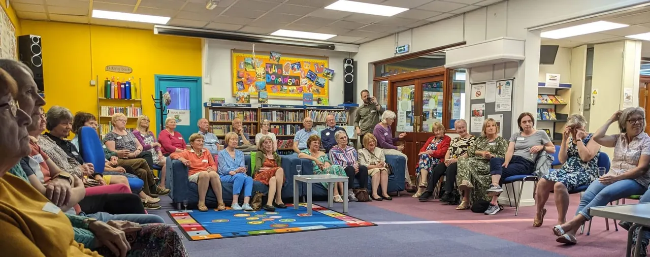 A group of older adults seated in a circle in a brightly decorated library room during an activity or gathering.