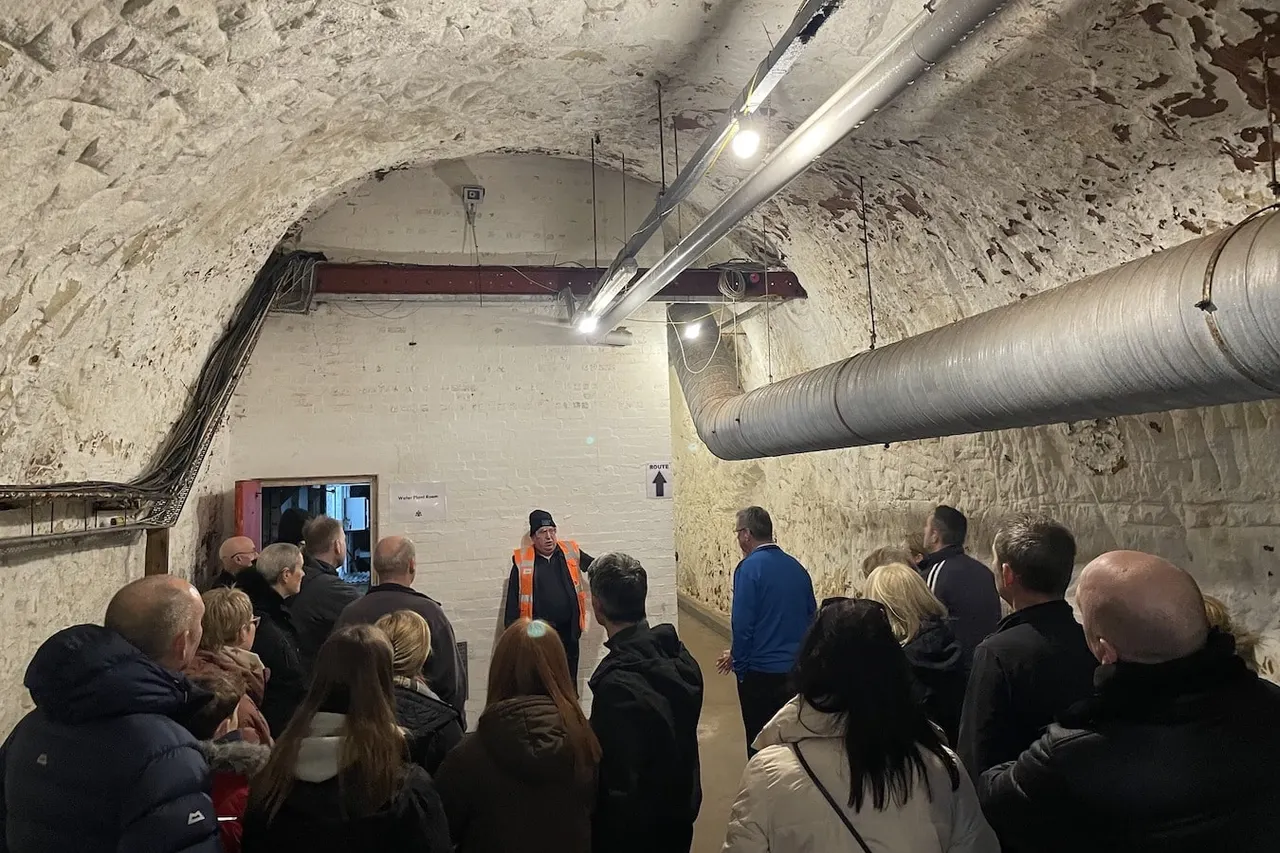 A tour guide in an orange safety vest speaks to a group of visitors inside a bare concrete underground tunnel with exposed pipes and industrial lighting.