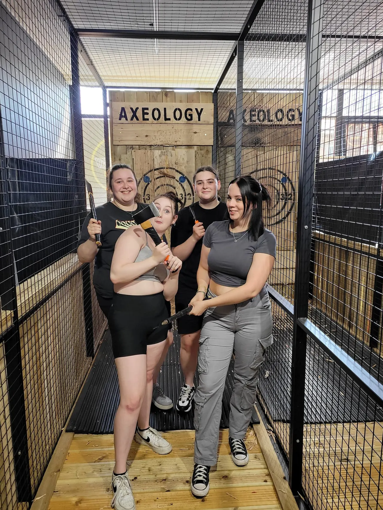 Four people holding axes inside a mesh-enclosed axe-throwing lane with a wooden target board marked "Axeology" at the end.