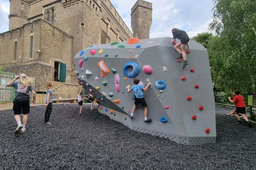 Children climbing a large indoor climbing wall with coloured holds in a castle courtyard.