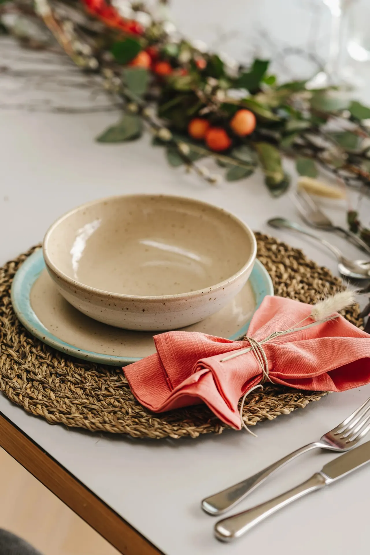 A formal place setting with stacked ceramic bowls on a woven placemat, coral napkin, and cutlery, decorated with autumn foliage and berries.
