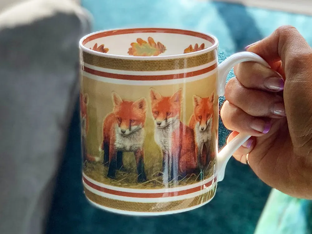 Someone's hand holding a mug decorated with images of foxes in a natural outdoor setting.