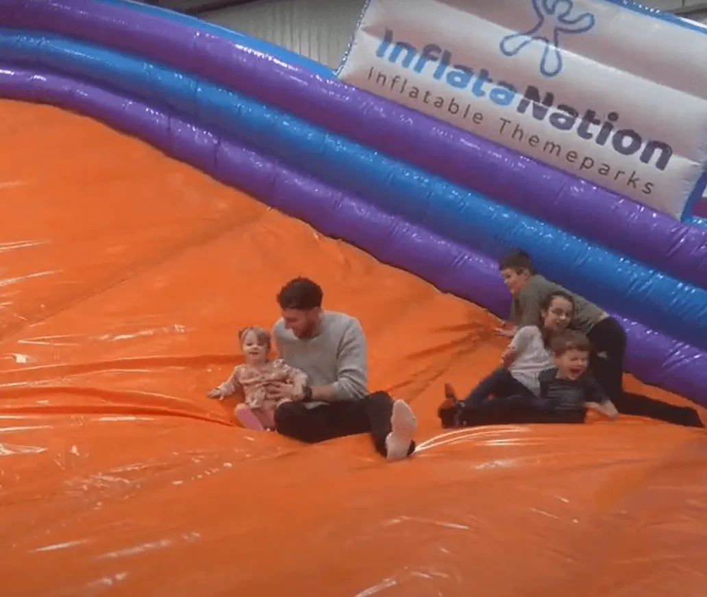 Adults and children playing together on a large orange inflatable structure inside an indoor theme park.