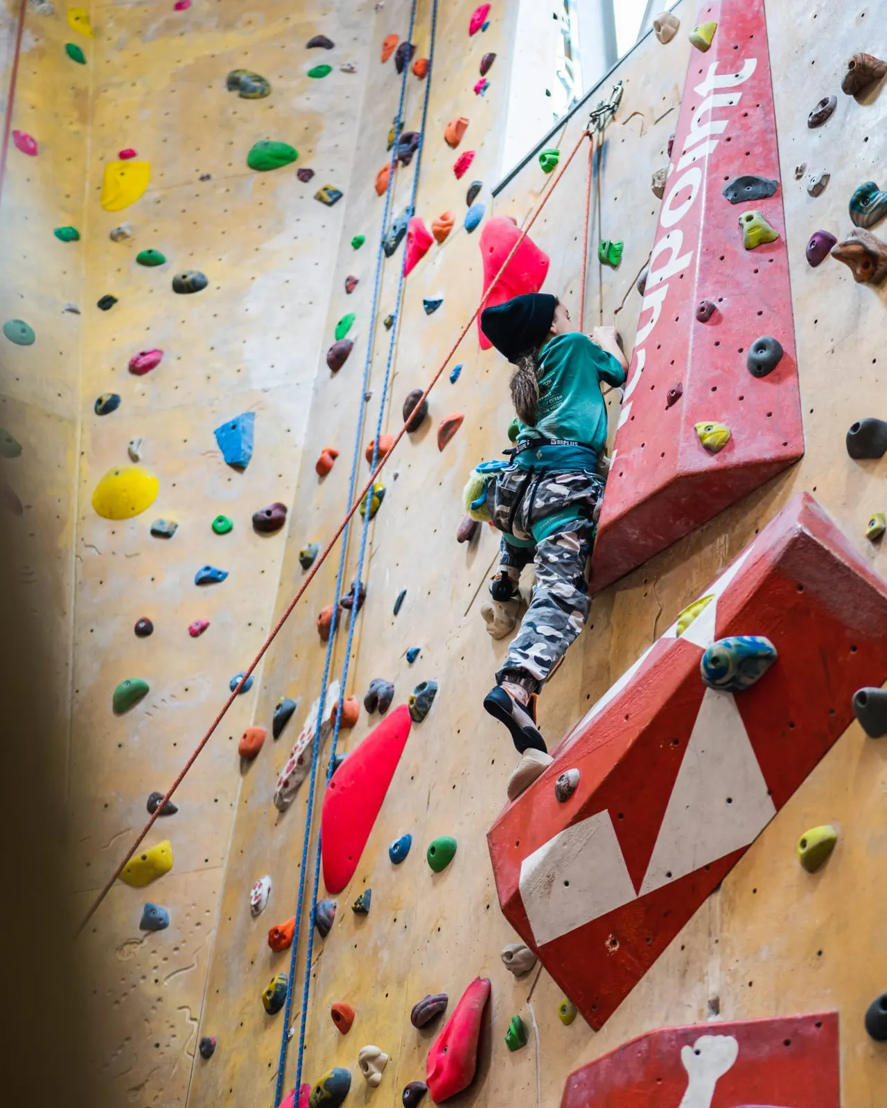 Child in pink helmet climbing an indoor rock wall with coloured holds, secured by rope.