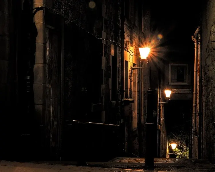 Narrow cobbled alleyway at night, lit by street lamps, with historic stone buildings on either side.