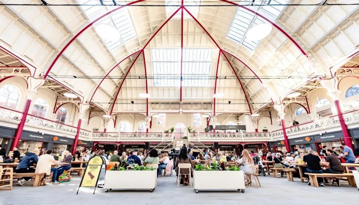 Bustling indoor market hall with high ornate ceiling, red ironwork, seated diners at wooden tables, and vendor stalls.