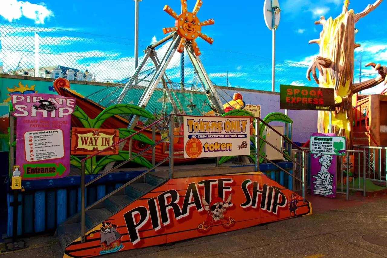 Colourful pirate-themed fairground ride entrance with spinning ship attraction, signage, and tropical decorations under blue sky.